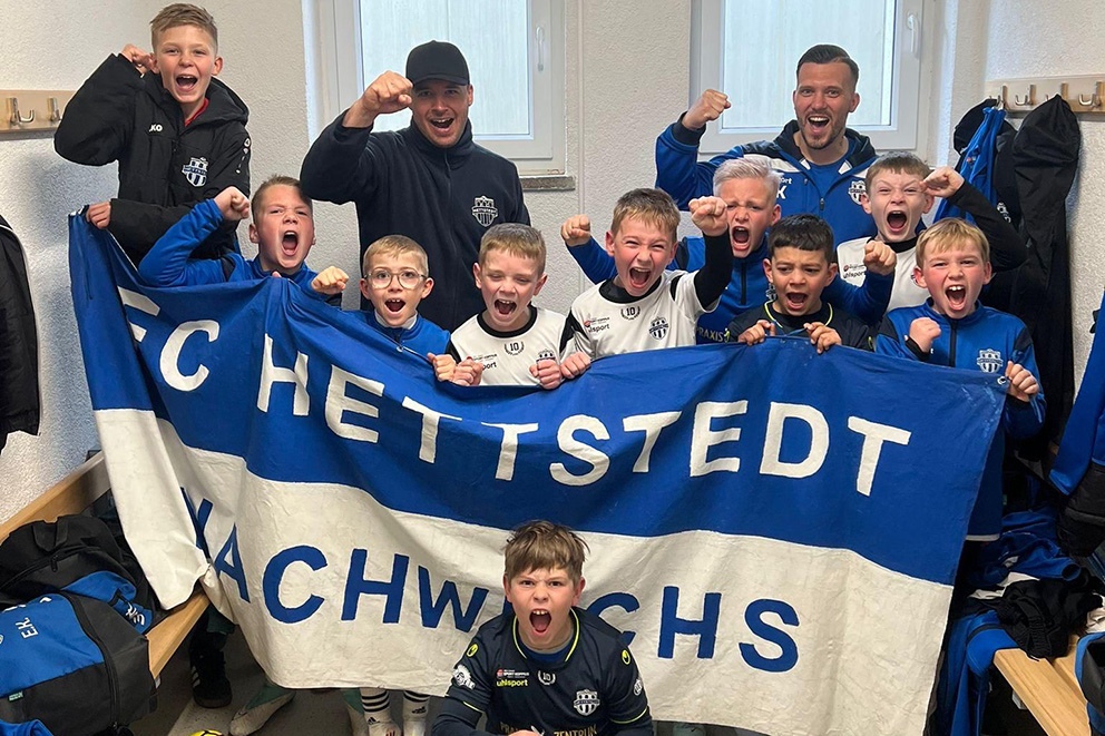 Jubelnde Nachwuchsspieler des FC Hettstedt mit Trainern und blau-weißer Vereinsfahne in der Kabine